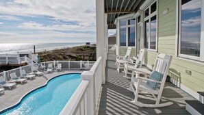 Et poolområde med udsigt til havet, hvide gyngestole og en overdækket terrasse.