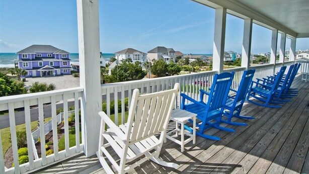 A covered porch with white and blue rocking chairs, a view of the ocean, and houses in the background.