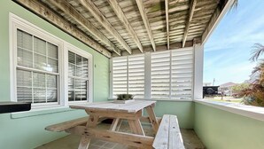 A covered patio with a wooden table and benches, large windows with blinds, and a view of the neighborhood. - Beach Life Down, Oceanview Duplex in Atlantic Beach, NC! (Atlantic Beach)