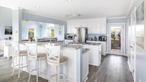 A modern kitchen with white cabinetry, a marble island, and a dining area with bar stools. - Beach Thyme, Oceanfront House in Indian Beach, NC! (Indian Beach)