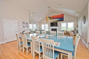 A spacious dining area with a blue table and white chairs, a ceiling fan, and a painting above the fireplace. - Beached, Almost Oceanfront House in Emerald Isle, NC! (Emerald Isle)