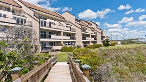 A multi-story apartment building with balconies, a wooden walkway, and a clear sky. - Beachwalk 112, Oceanfront Condo in Pine Knoll Shores, NC! (Pine Knoll Shores)