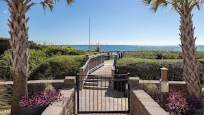 A wooden boardwalk leading to a beach with palm trees and a clear sky. - Beachwalk 112, Oceanfront Condo in Pine Knoll Shores, NC! (Pine Knoll Shores)