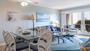 A living room with a glass coffee table, wicker chairs, a blue sofa, and a ceiling fan. - Beachwalk 112, Oceanfront Condo in Pine Knoll Shores, NC! (Pine Knoll Shores)