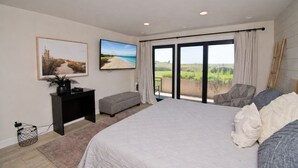 A bedroom with a large bed, a TV, a desk, and a view of a vineyard through the windows. - Beachwalk 116, Oceanfront Condo in Pine Knoll Shores, NC! (Pine Knoll Shores)