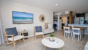A modern living room with a large TV, a wooden dining table, and a sofa. - Beachwalk 116, Oceanfront Condo in Pine Knoll Shores, NC! (Pine Knoll Shores)