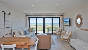 A spacious living area with a wooden dining table, a sofa, and a balcony with a view of the beach. - Beachwalk 116, Oceanfront Condo in Pine Knoll Shores, NC! (Pine Knoll Shores)