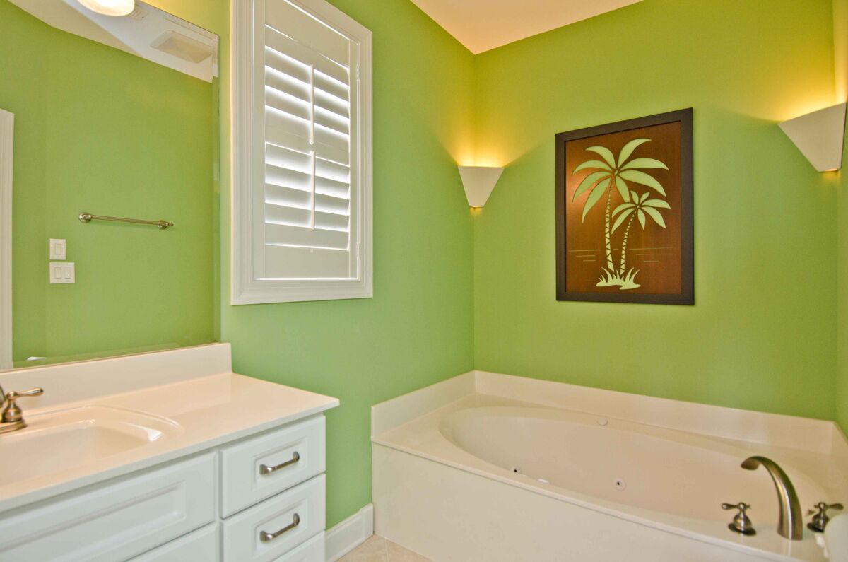 Un baño con paredes verdes, un lavamanos blanco, una bañera y un cuadro con palmeras.