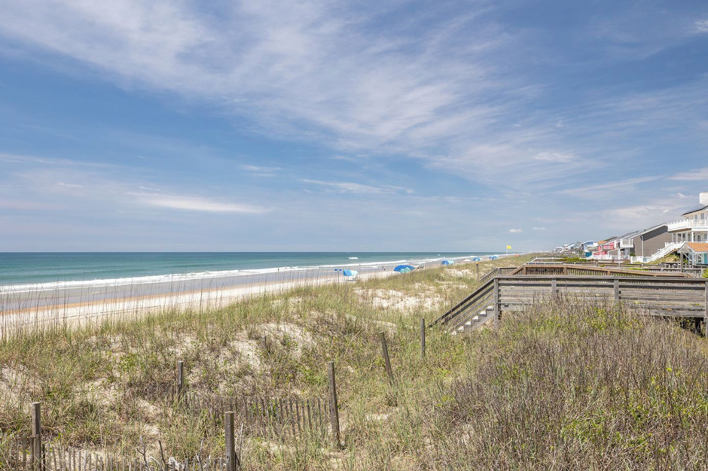 A beach view with a wooden boardwalk, beach umbrellas, and houses in the background.