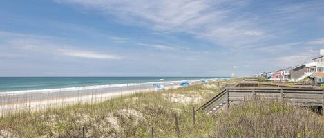 A beach view with a wooden boardwalk, beach umbrellas, and houses in the background.