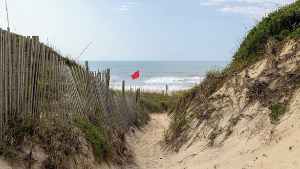 A sandy beach path leading to the ocean, with a wooden fence on one side and a red flag on the other.