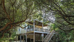 A two-story wooden cabin with a balcony and a staircase, surrounded by dense trees.