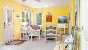 A room with yellow walls, a ceiling fan, a television, and a window with a view of greenery.