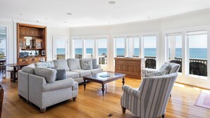 A spacious living room with a large sofa, armchairs, and a coffee table. The room has large windows with ocean views. - Bliss By The Beach, Oceanfront House in Emerald Isle, NC! (Emerald Isle)