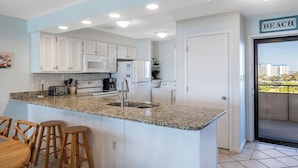 A kitchen with white cabinets, a granite countertop, a sink, a toaster, a coffee maker, and a refrigerator. There is a door with a 'BEACH' sign above it.