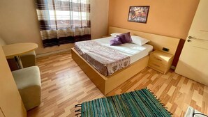 A bedroom with a bed, a small table, a chair, a window with curtains, a painting on the wall, and a door. - Algo Hotel (Tirana)