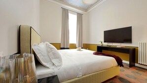 Quarto de hotel com cama, televisão de tela plana, mesa de cabeceira, espelho e janela com cortinas.