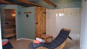Una habitación con una silla de masaje de madera, una mesita pequeña de madera y una cabina de ducha de vidrio.