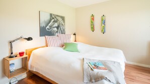 Quarto bem arrumado com cama, almofadas, mesa de cabeceira com abajur, pequena estante com um livro e um quadro na parede.
