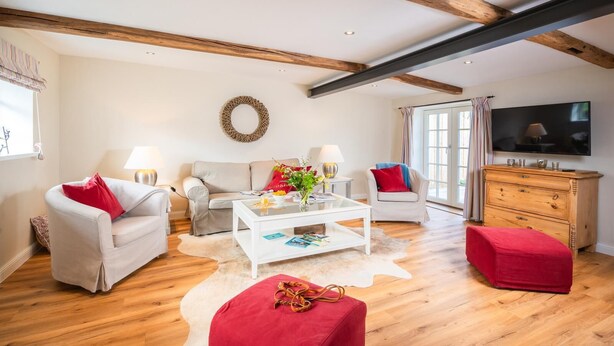 A spacious living room with a high ceiling, wooden beams, a white sofa, a red ottoman, a coffee table, and a television.