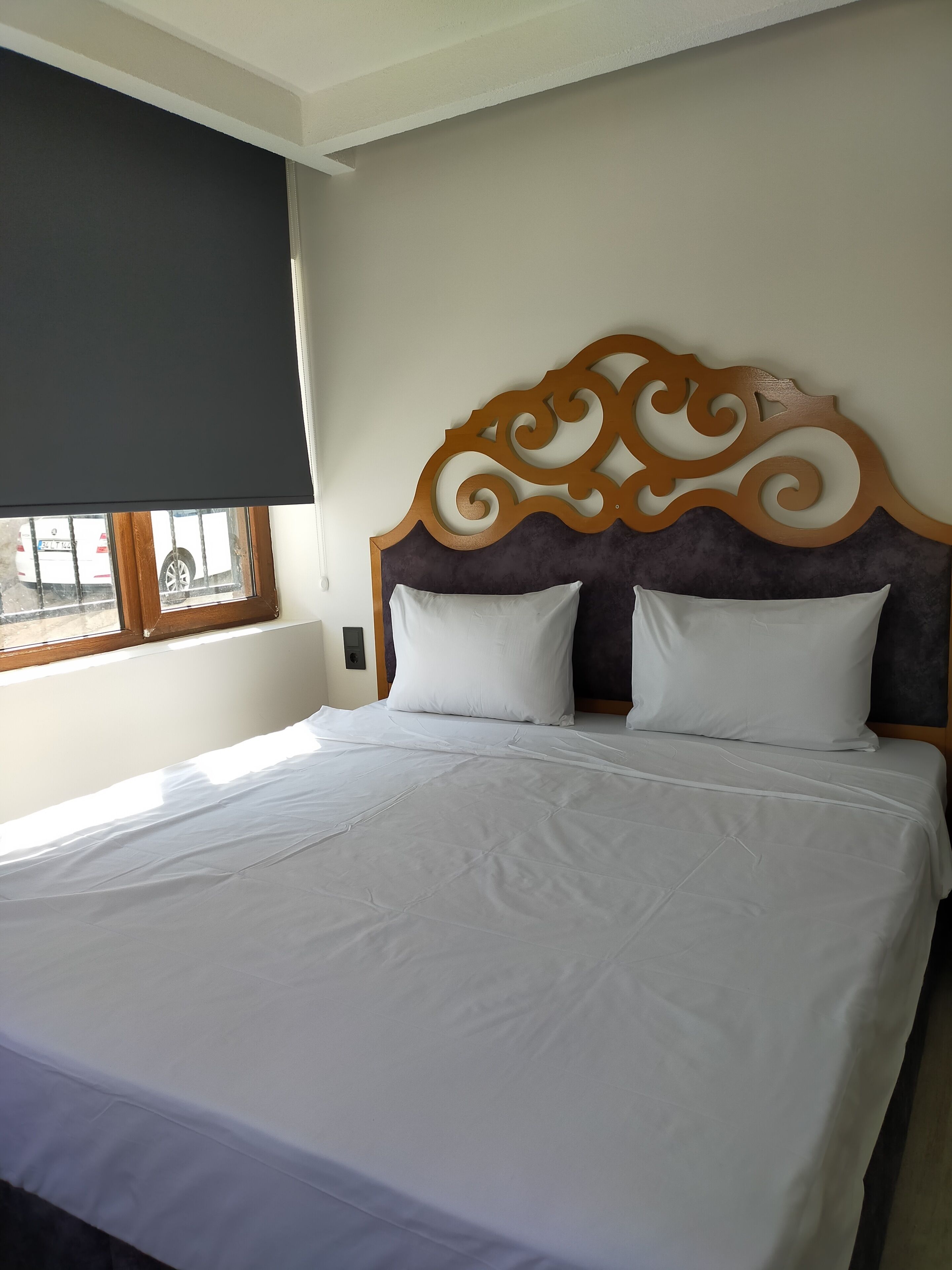 Una cama bien hecha con cabecera de madera, ropa de cama blanca y una ventana que da a la vista de un coche estacionado afuera.