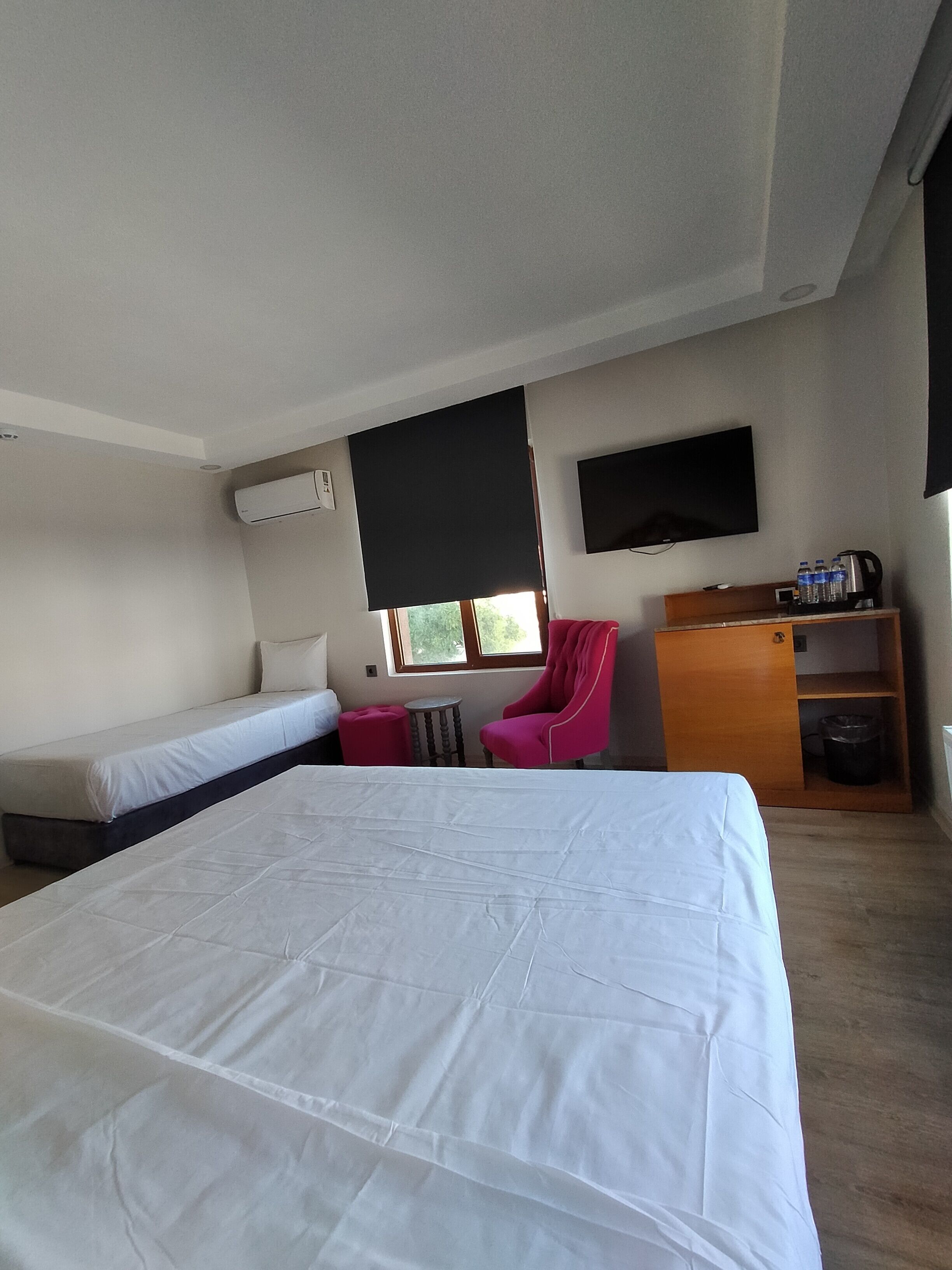 Habitación de hotel con una cama grande, otra más pequeña, televisor montado en la pared, una silla, un escritorio con cafetera y una ventana con vistas a zonas verdes.