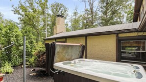 A hot tub in a backyard with a wooden deck, surrounded by trees and a house with a chimney. - Relaxing & Rejuvenating Retreat w/Hot tub, Pool, Sauna, & Games for family+pets (Berkeley Springs)