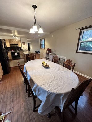 Un comedor con una gran mesa redonda cubierta con un mantel blanco, rodeada de sillas de madera. Un frutero está colocado sobre la mesa. El cuarto tiene piso de madera y un accesorio de iluminación en el techo.