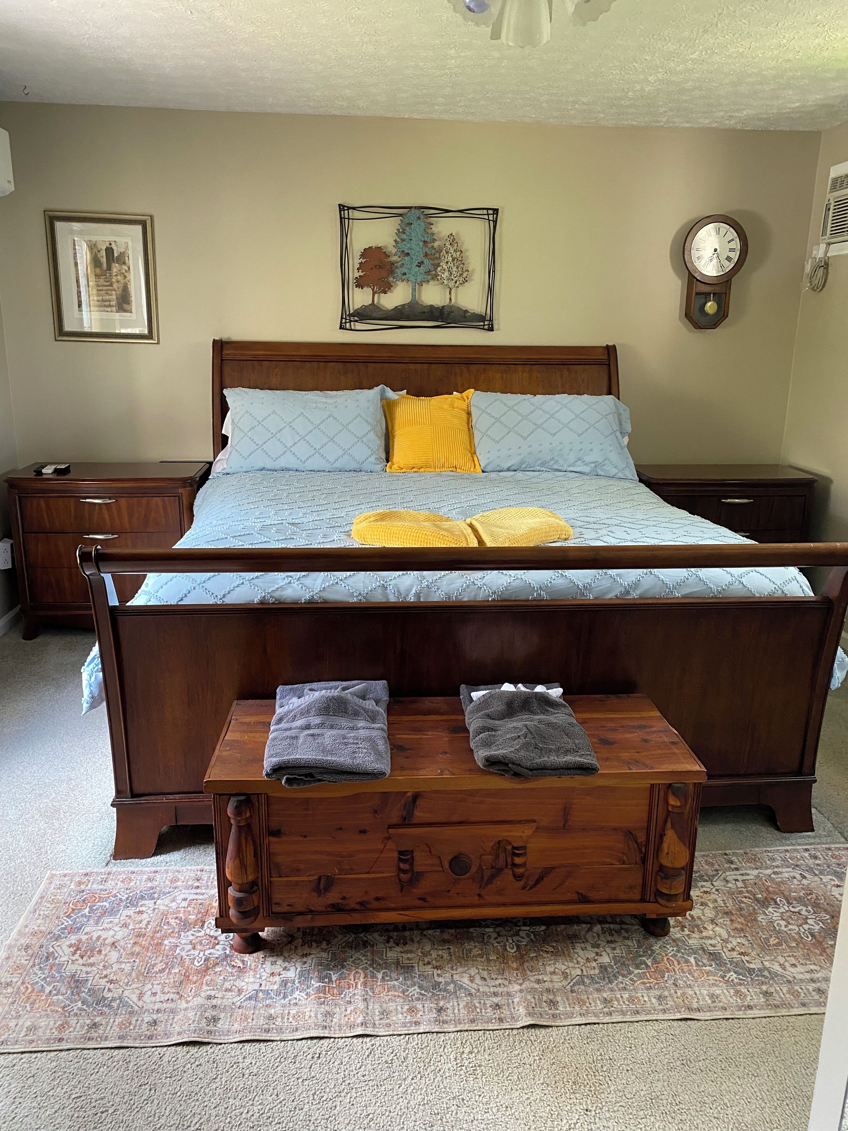 Ein Schlafzimmer mit einem hölzernen Bett, zwei Nachttischen, einer Holzbank mit Handtüchern, einem gemusterten Teppich und einer Uhr an der Wand.