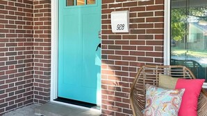 A turquoise door with a number 928 sign, a wicker chair with a colorful cushion, and a black doormat. - The Vandalia House - Midcentury Rodeo Meets Midtown, University of Tulsa, and Expo Center (Tulsa)