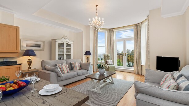 A spacious living room with a grey sofa, a wooden coffee table, and a chandelier. There is a television on the right and a cabinet on the left. - Bryn Y Mor - Swimming Pool and  Sea Views (Tenby)