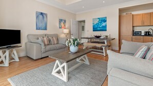 A modern living room with a sofa, coffee table, and TV. There is a kitchen area with wooden cabinets and a dining table. - Bryn Y Mor - Swimming Pool and  Sea Views (Tenby)