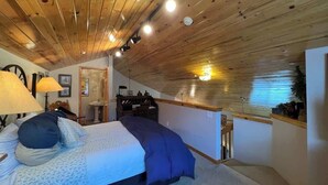A bedroom with a wooden ceiling, a bed with blue bedding, a desk with a lamp, and a small bathroom area. - Swan River Condo w/ Hot Tub & Fireplace (Breckenridge)