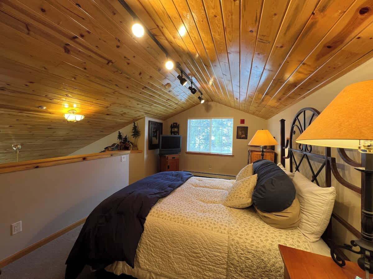 Une chambre confortable avec un plafond en bois, un lit recouvert d’une courtepointe, deux lampes de chevet et une fenêtre avec des stores.