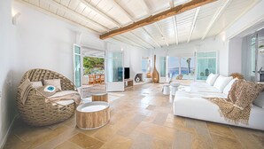 A spacious living area with a high ceiling, a large sofa set, a wicker chair, and a view of the sea through large windows. - Sanarte  Villas Mykonos (Mykonos)