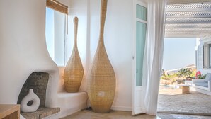 A modern interior with two tall, woven vases, a stone fireplace, and a view of an outdoor area with a pool and lounge chairs. - Sanarte  Villas Mykonos (Mykonos)