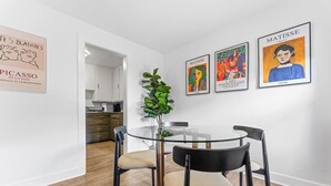 Ein moderner Essbereich mit einem Glastisch und schwarzen Stühlen, einer Topfpflanze und drei gerahmten Kunstwerken an der Wand.