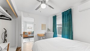 Ein Schlafzimmer mit einem Bett, einem Schreibtisch und einem Stuhl. Es gibt einen Deckenventilator, ein Fenster mit Vorhängen und einen Blick ins Freie.