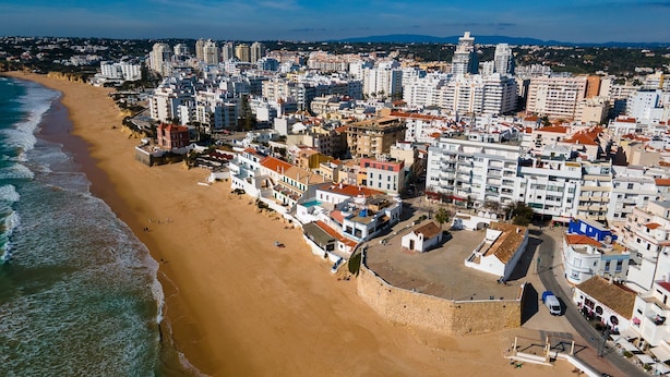 Vista aérea de una ciudad costera con playa, edificios y una muralla fortificada.