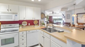 Una cocina moderna con armarios blancos, un microondas, un horno y un lavavajillas. Se aprecia una pared de acento roja, un ventanal grande y un área de comedor al fondo.