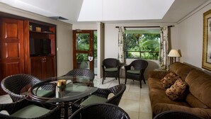 Une pièce avec une table en verre, des chaises en osier, un canapé marron, un ventilateur de plafond et une vue sur la verdure par une fenêtre.