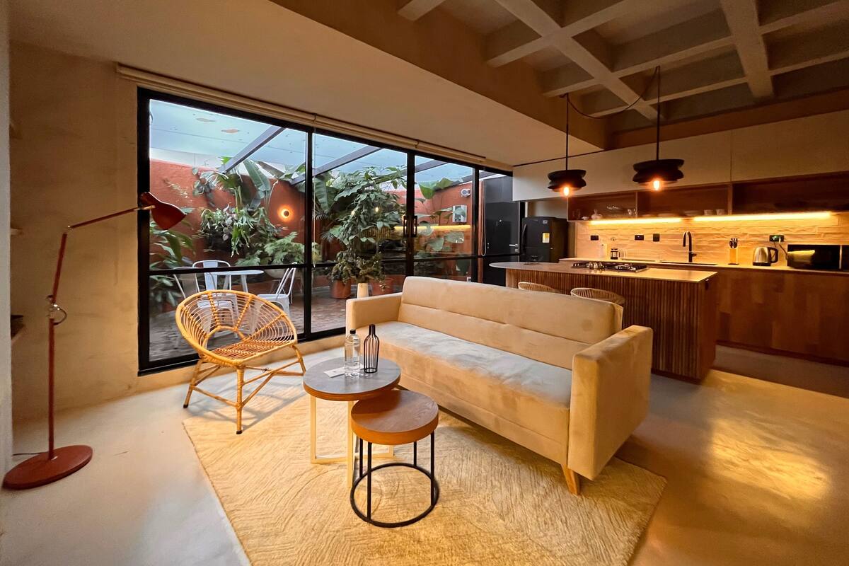 Una sala de estar moderna con un sofá, dos sillones, una mesita y un amplio ventanal corredera que da a un balcón con plantas.