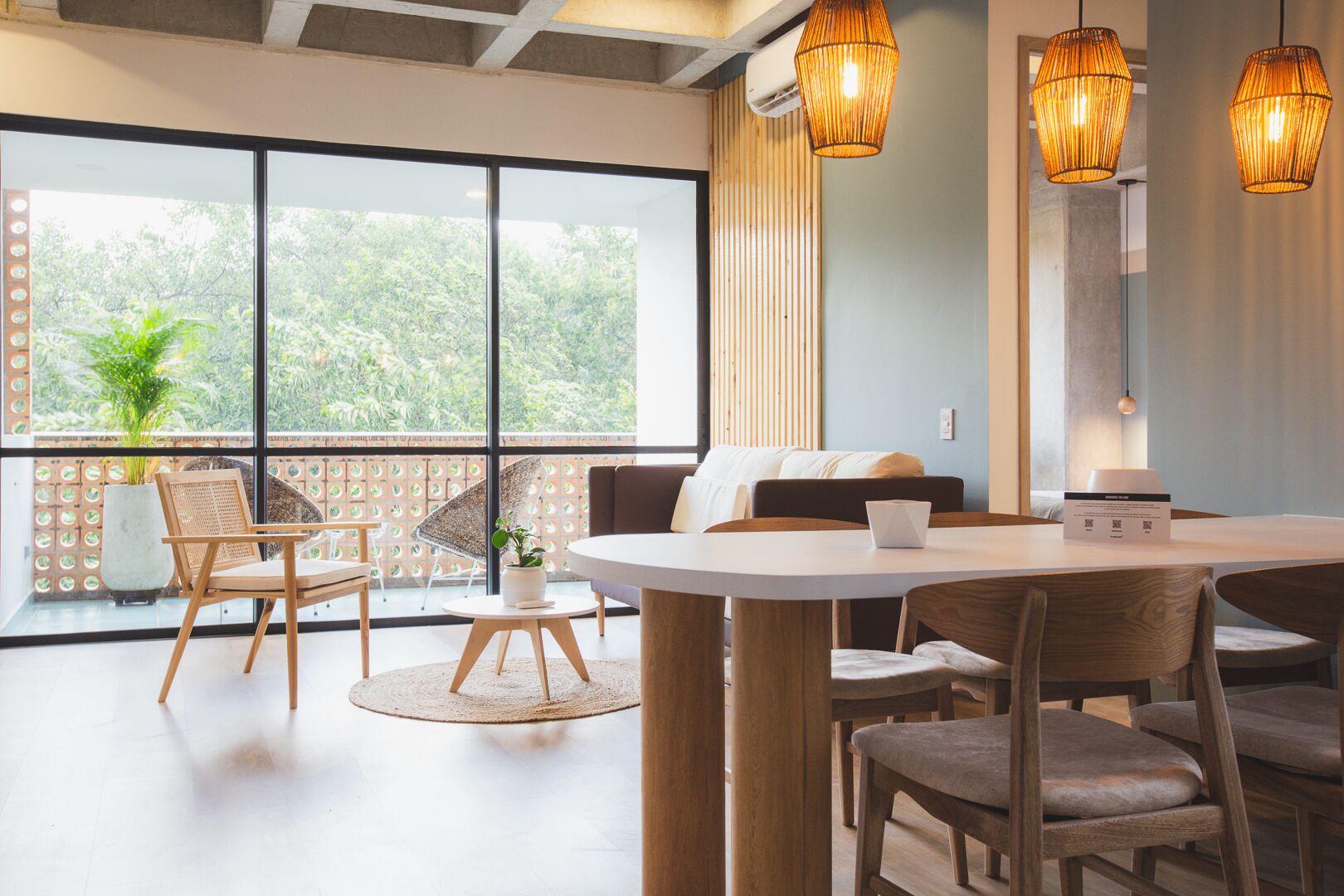 A modern room with a round dining table, wooden chairs, a sofa, and a large window with a view of greenery.