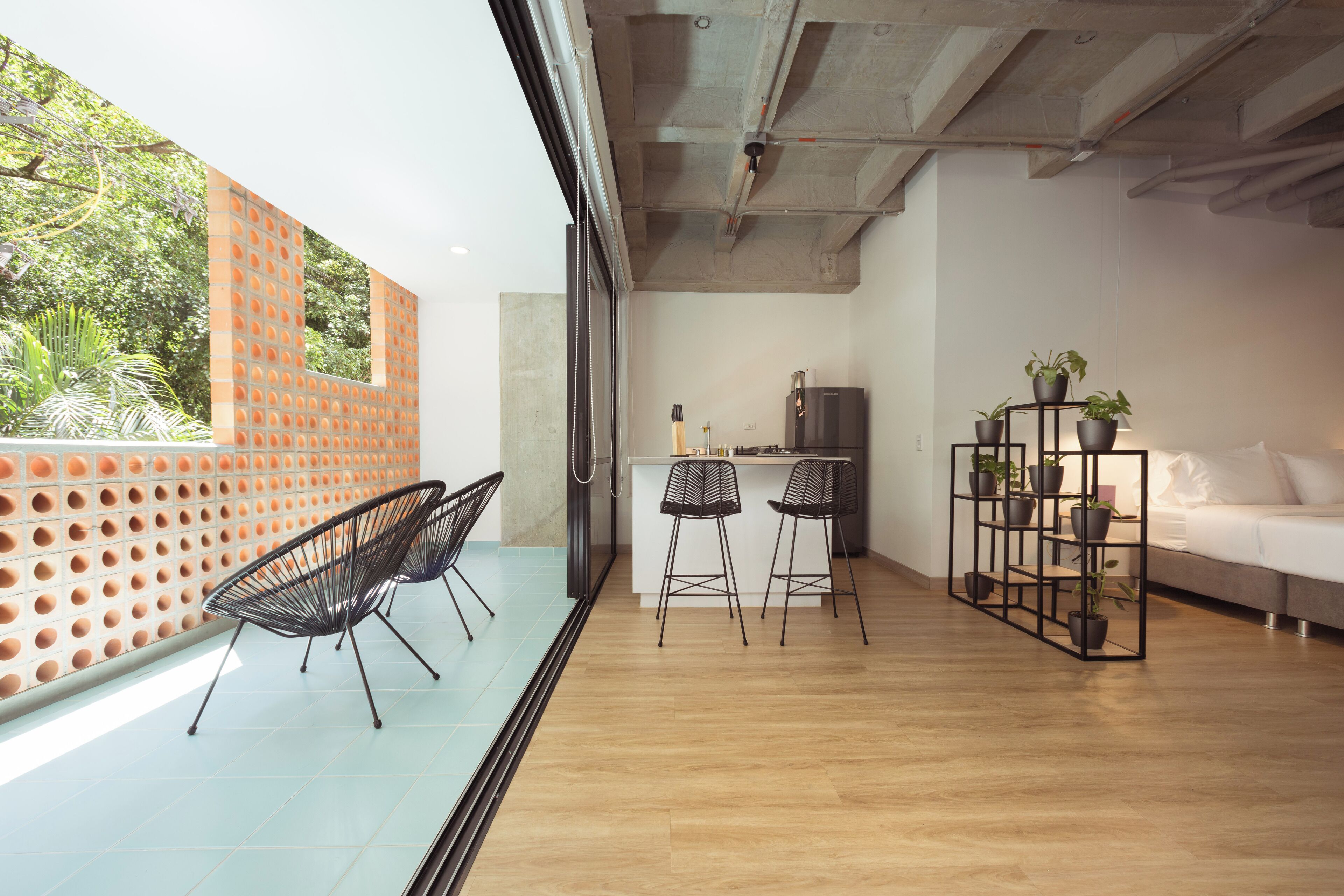 Un interior moderno con una zona de cocina, dos taburetes, un sofá y una estantería con plantas.