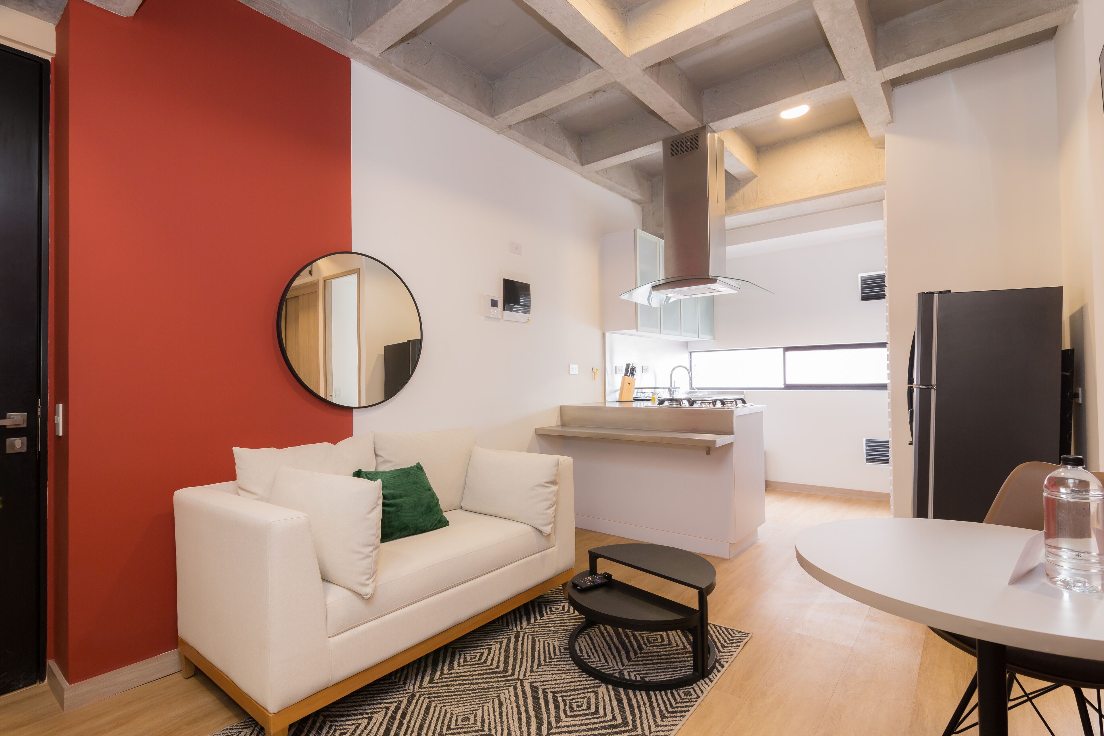 A modern living room with a white sofa, a round mirror, and a small coffee table. A kitchen area is visible in the background with a refrigerator and a sink.