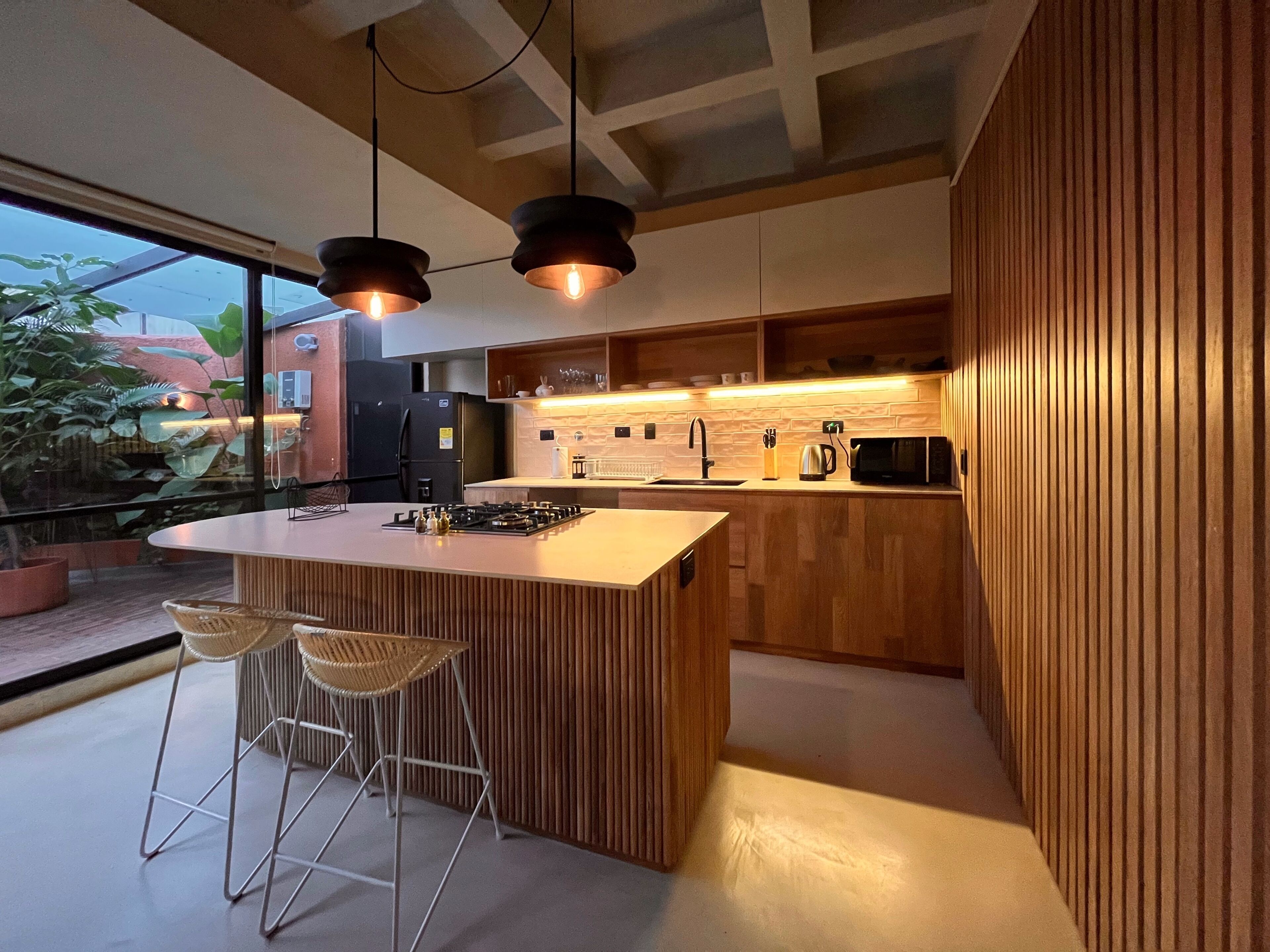A modern kitchen with a central island, bar stools, wooden cabinetry, and pendant lighting.