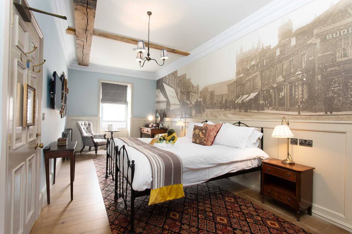 A bedroom with a large bed, a nightstand, a chair, a desk, and a wall-mounted TV.