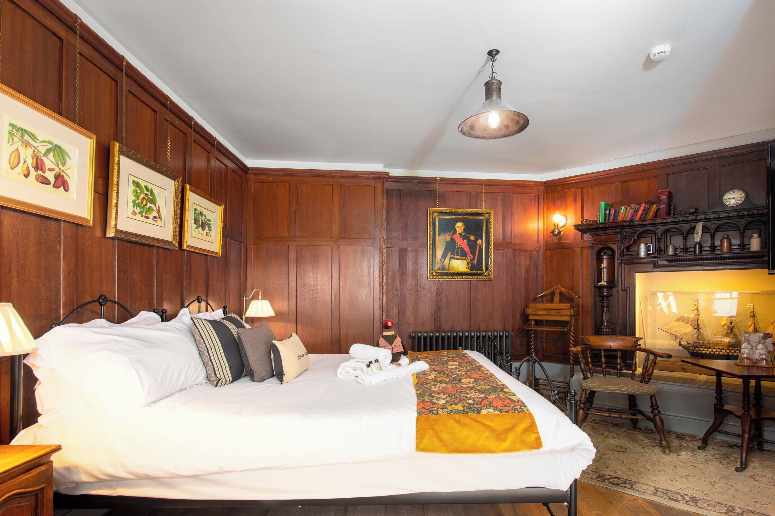 A hotel room with a large bed, wooden paneling, framed pictures on the wall, a desk with a lamp, and a ship model in a glass case.