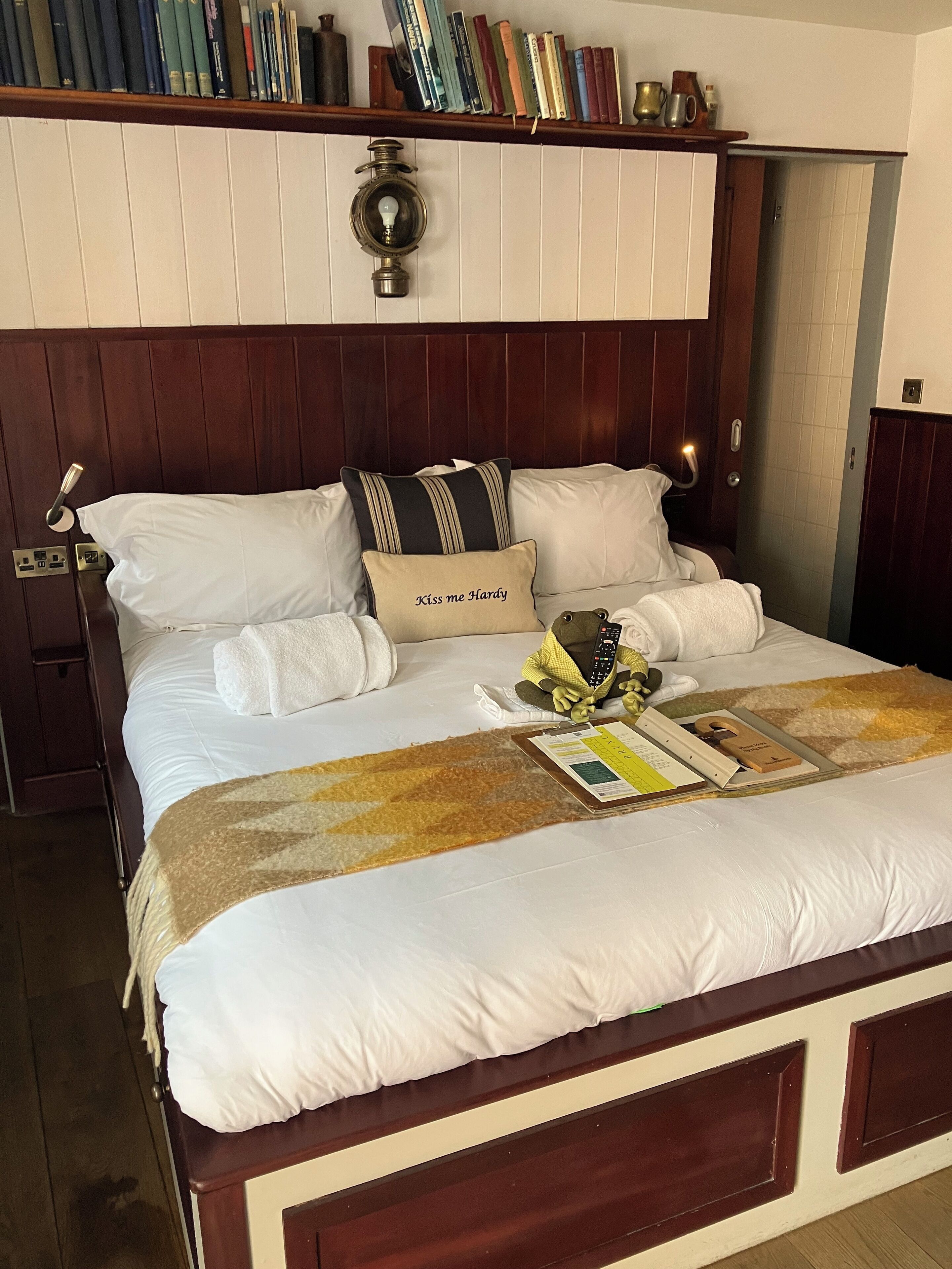 Una cama bien hecha con edredón blanco y almohadas, cabecera de madera, lámpara de noche y estante con libros y objetos decorativos.