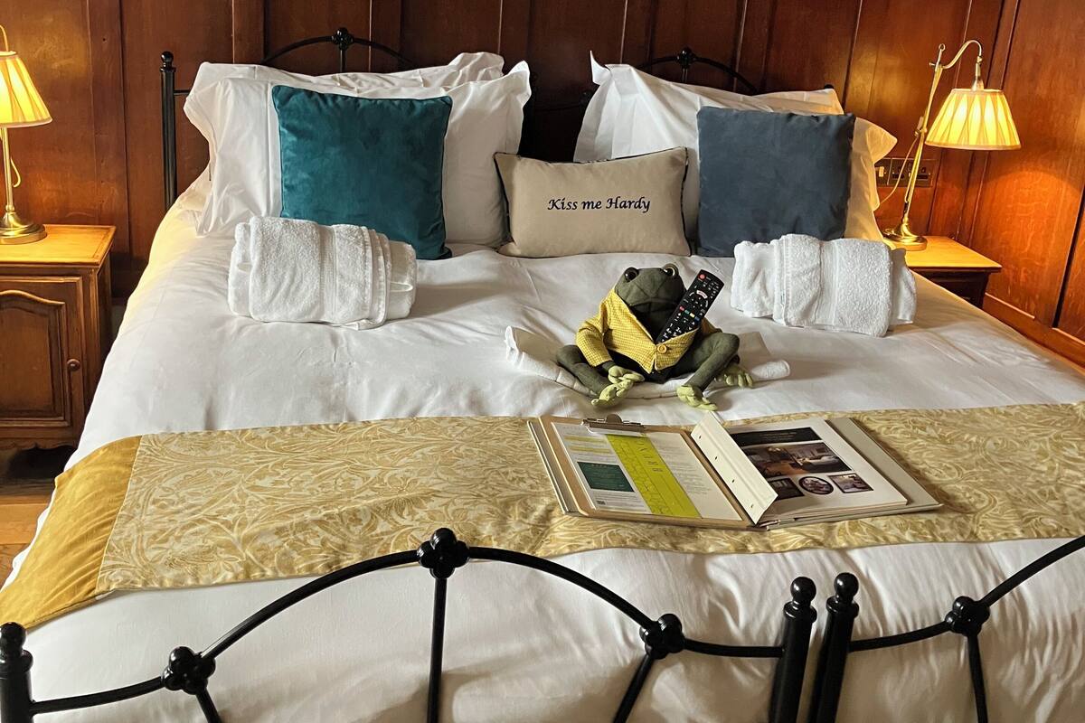 A neatly made bed with white linens, multiple pillows, and a bedside table with a lamp. There are framed botanical illustrations on the wall above the bed.